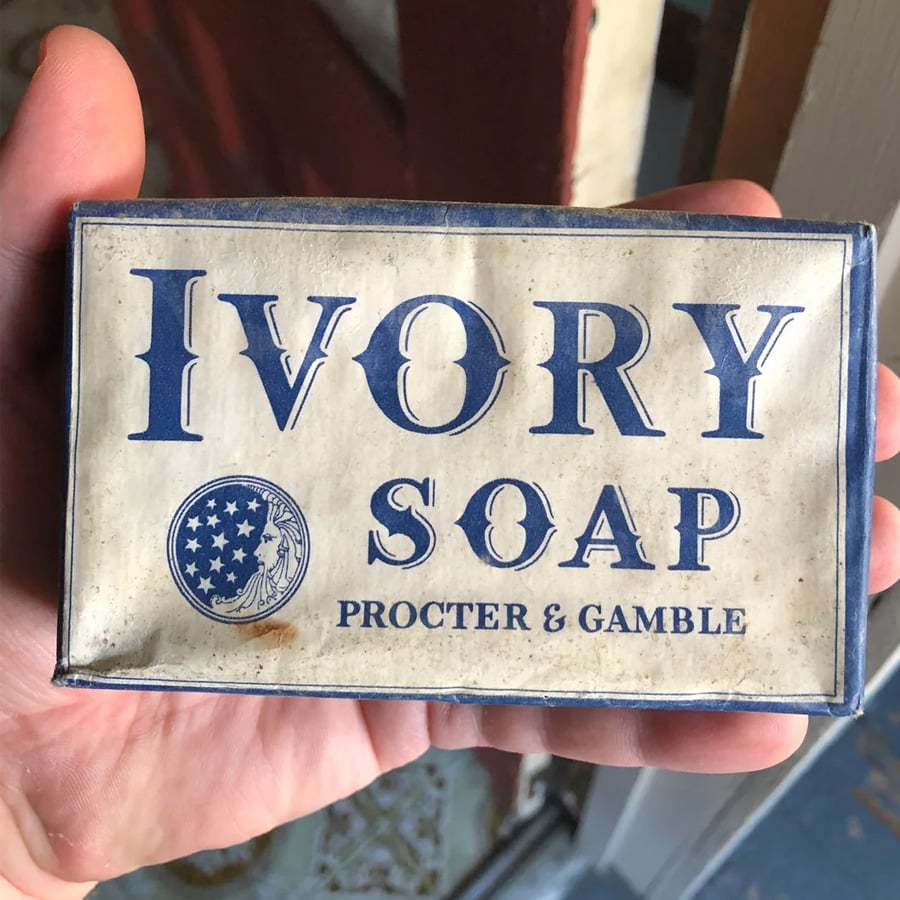 A hand holds a vintage bar of Ivory Soap by Procter & Gamble. The white and blue packaging features a star-spangled circular logo and bold letters. The box appears old with some stains and wear.