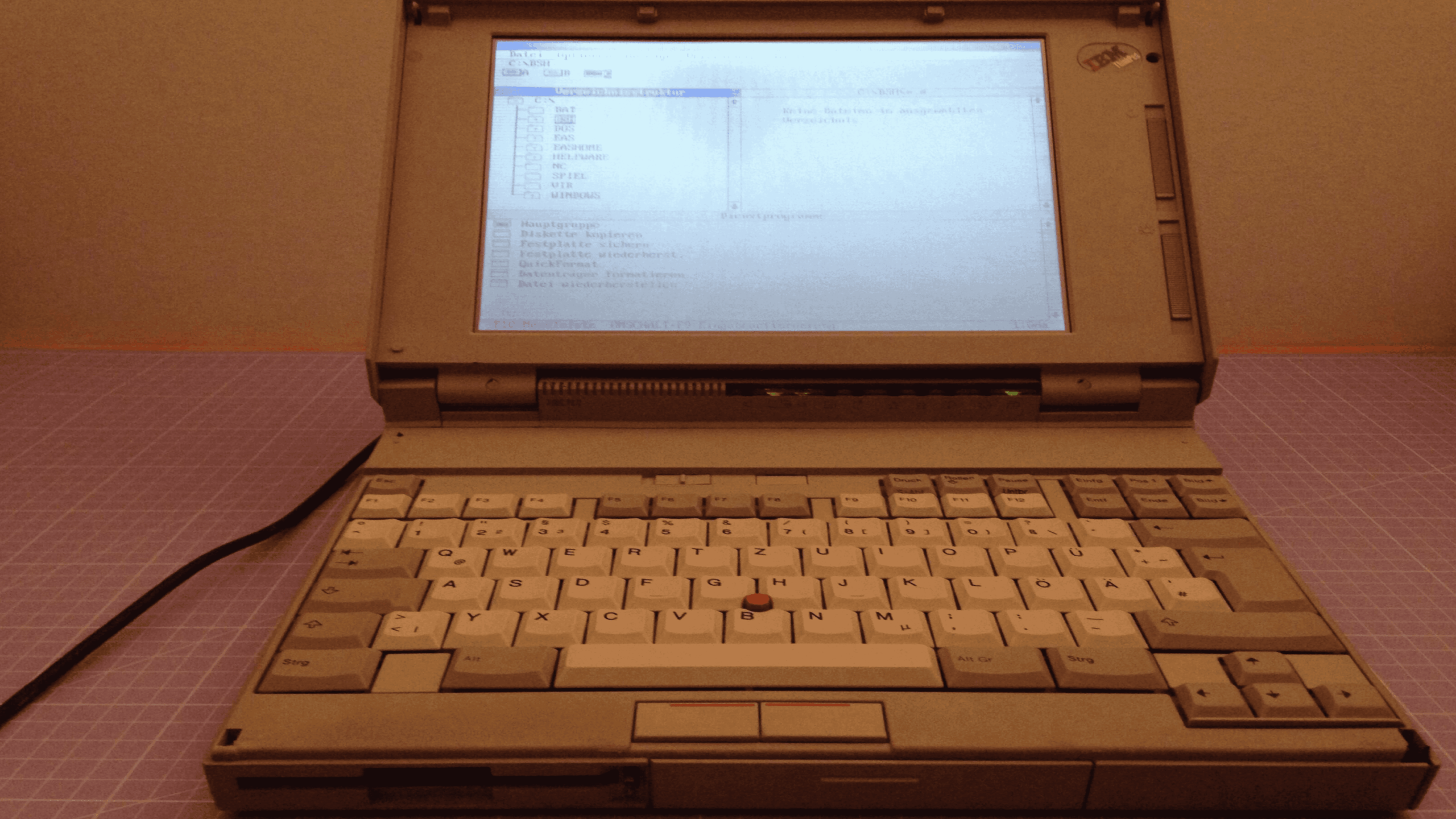 An old gray laptop with a screen displaying text, a compact keyboard, and a red pointing stick in the center, sitting on a purple grid-patterned surface.