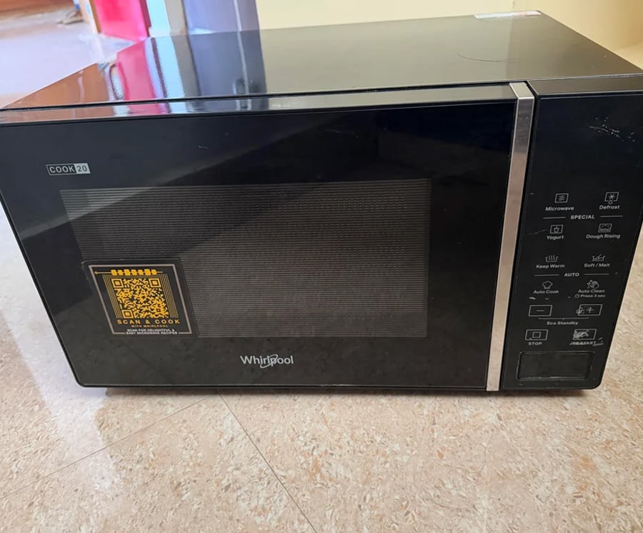 A black Whirlpool microwave oven with a control panel on the right side and a QR code sticker on the door, sitting on a light-colored floor.
