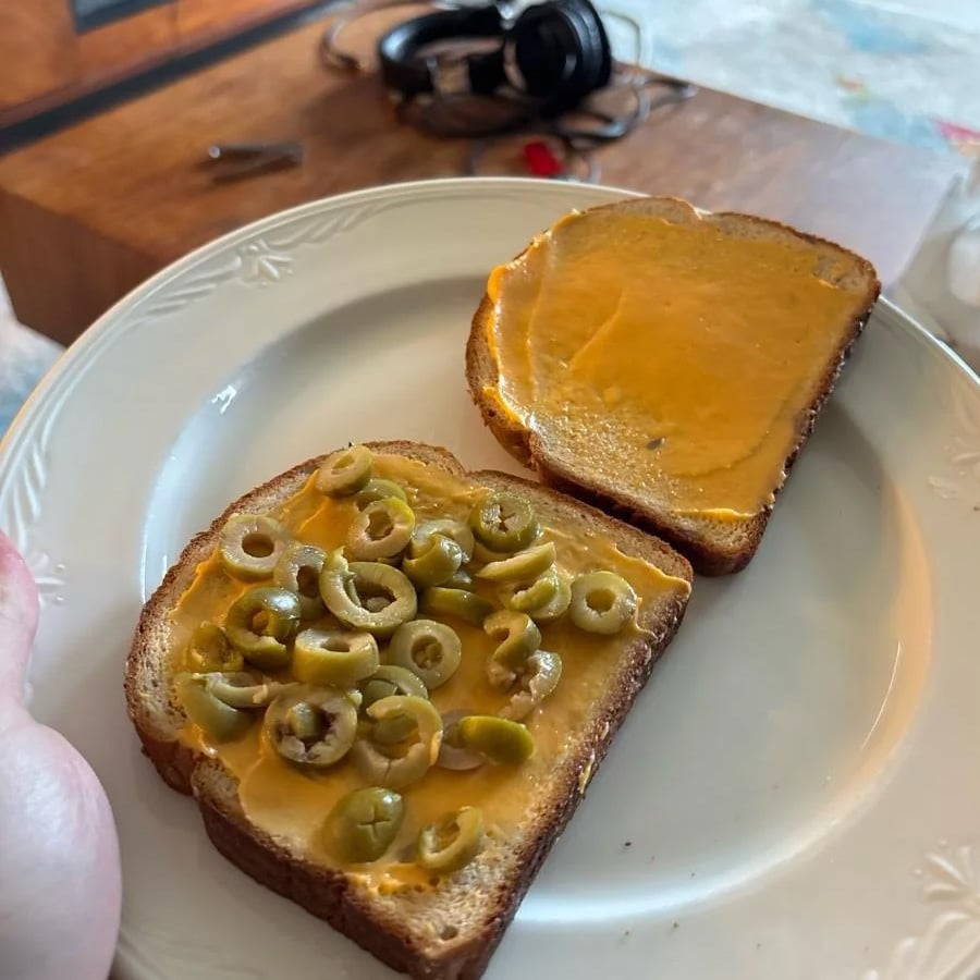 Two slices of bread with melted cheese sit on a white plate; one slice is topped with sliced green olives. A wooden table and headphones are visible in the background.