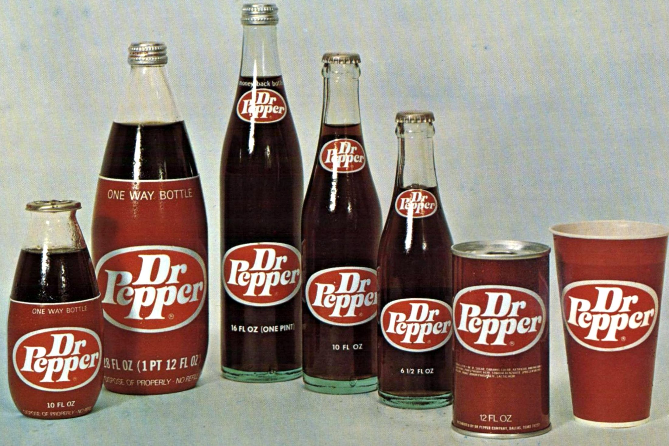 Six different Dr Pepper containers—glass bottles, cans, and a paper cup—are displayed in a row on a neutral surface, showcasing various sizes and the classic red and white Dr Pepper logo on each.