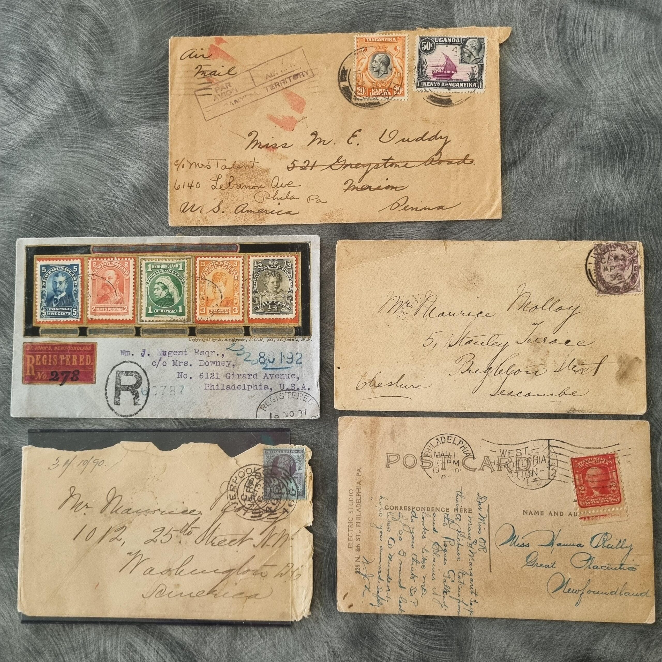 Six vintage envelopes and postcards with handwritten addresses and colorful, old postage stamps are displayed on a gray textured surface. Some are registered, and the letters show signs of age.