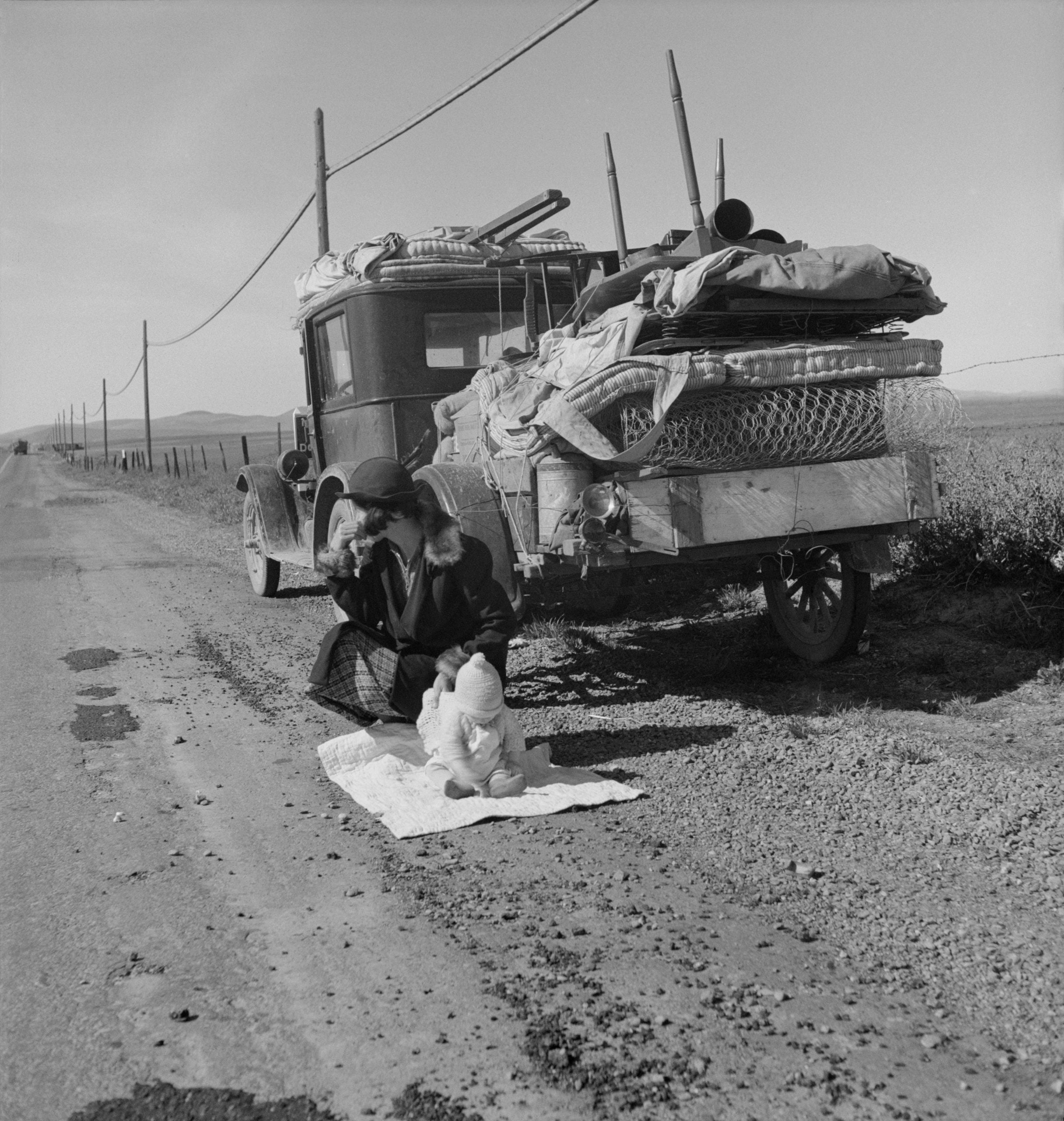 A woman and a baby sit on a blanket beside a heavily loaded old car on a rural road. Fields and power lines stretch into the distance under a clear sky.