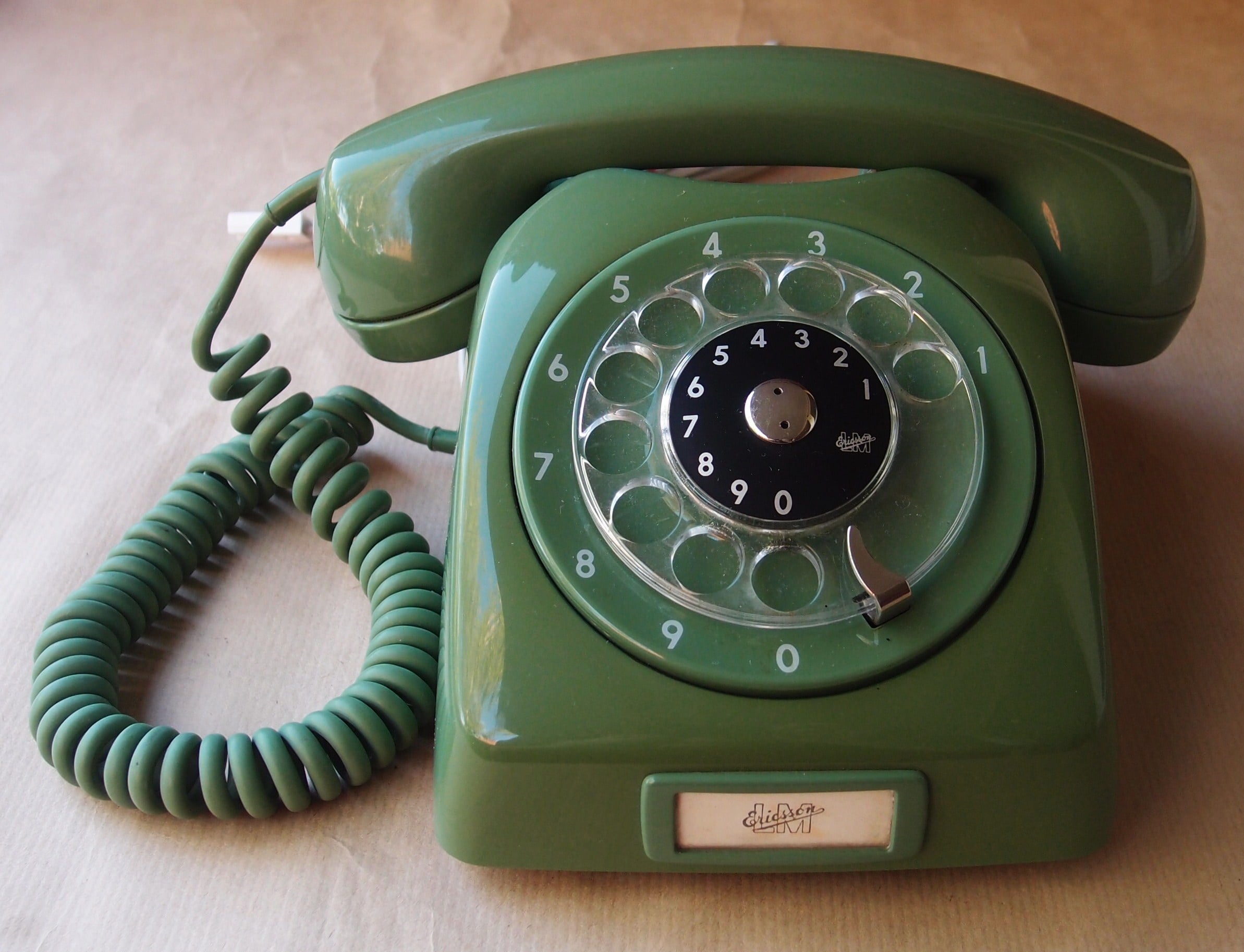 A vintage green rotary dial telephone with a coiled cord, sitting on a beige surface.