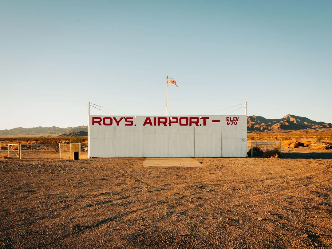 A white building with “ROY’S AIRPORT” painted in red stands in a desert landscape with mountains in the background. A small flag flies on top.