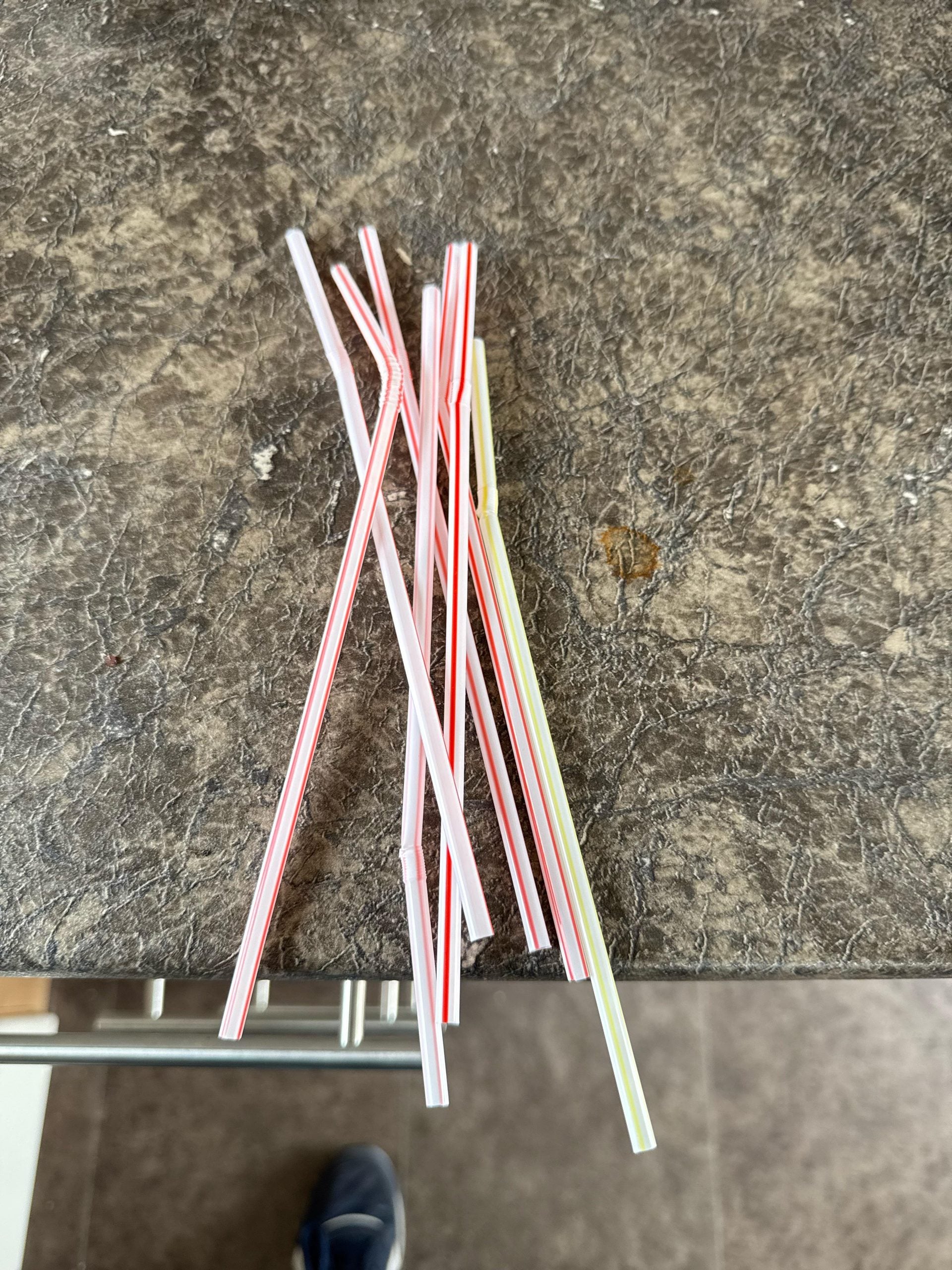 Several plastic drinking straws with red, green, and yellow stripes are scattered on a brown textured countertop. A partial shoe is visible at the bottom edge of the image.
