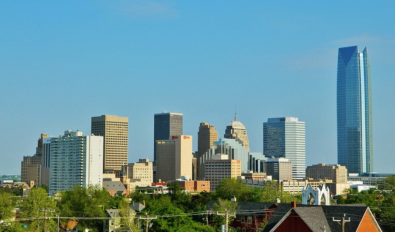 Oklahoma City skyline with various skyscrapers, including a tall glass building on the right, under a clear blue sky. Trees and smaller buildings appear in the foreground.