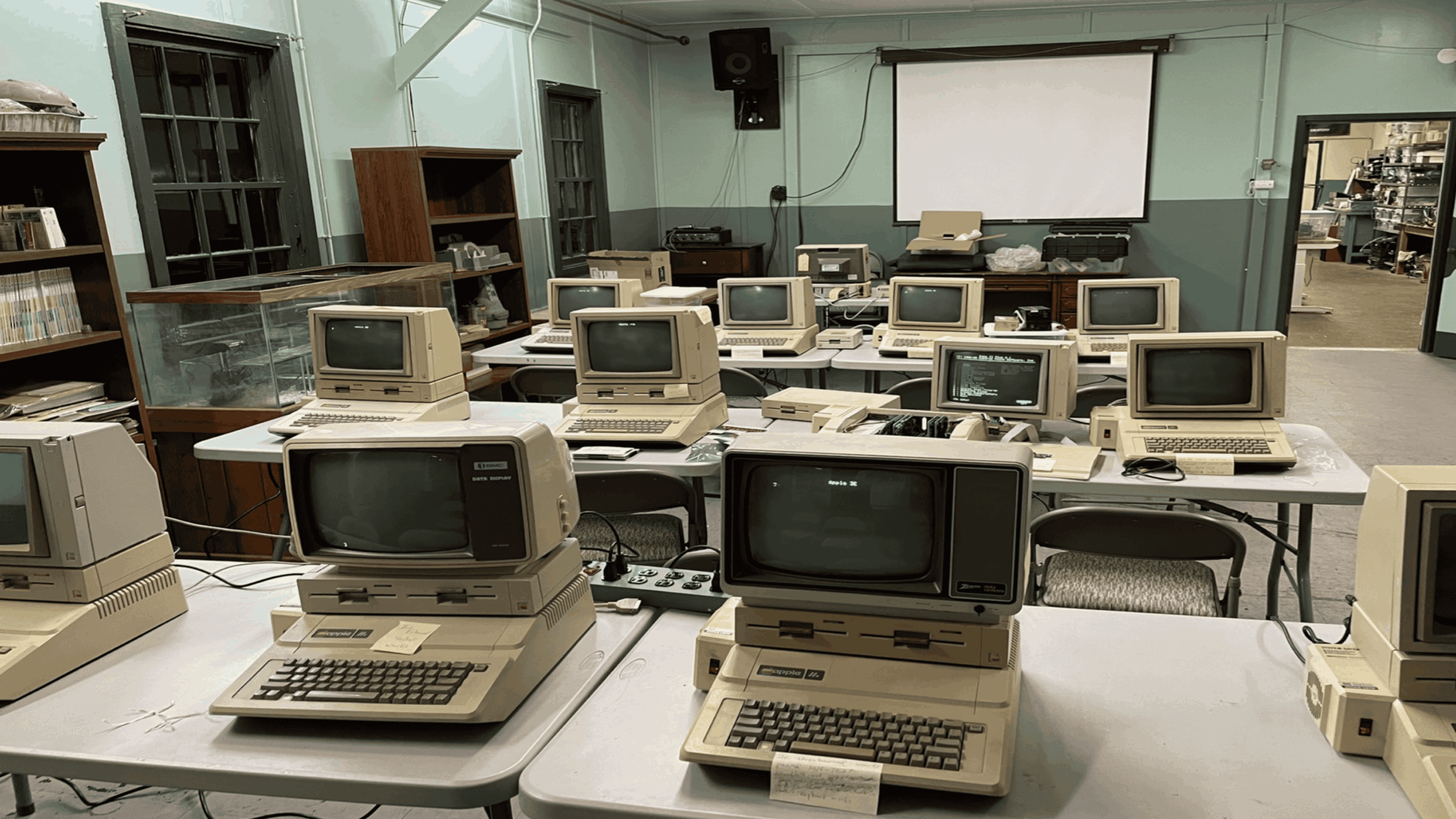 Vintage computer classroom with rows of old desktop computers on tables, CRT monitors, keyboards, and floppy drives in a room with green walls, shelves, and a projector screen.