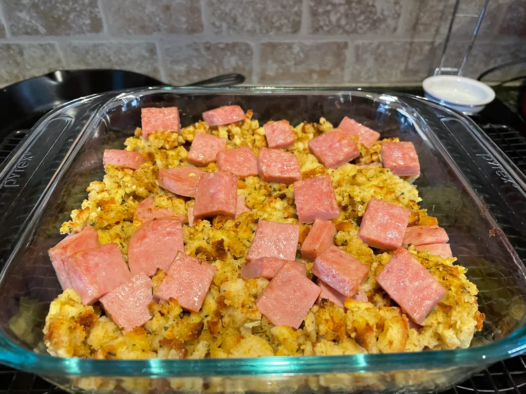 A glass baking dish filled with a casserole topped with cubed pink meat, possibly ham or spam, on a layer of seasoned bread or stuffing, with a kitchen counter background.
