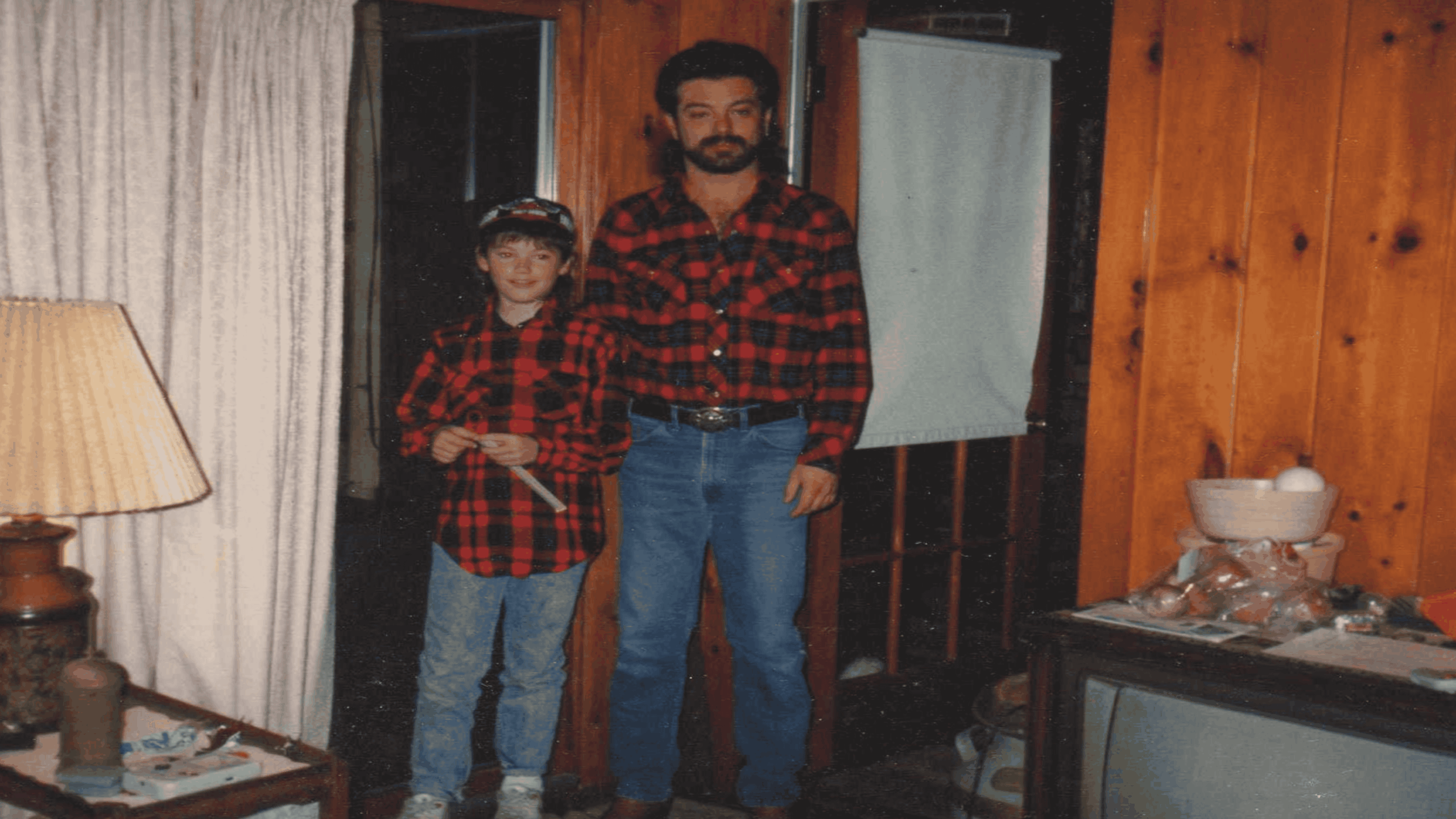 A man and a boy, both wearing matching red plaid shirts and jeans, stand side by side in a wood-paneled room next to a lamp and a TV.