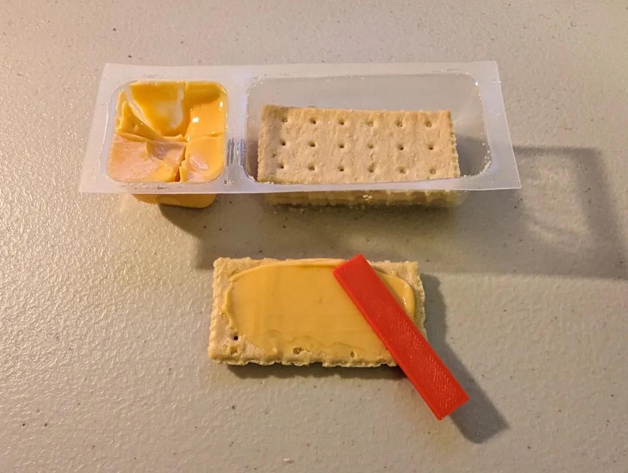 A snack pack with processed cheese, crackers, and a red plastic stick. One cracker is spread with cheese and the red stick is resting on top. The pack is on a beige surface.