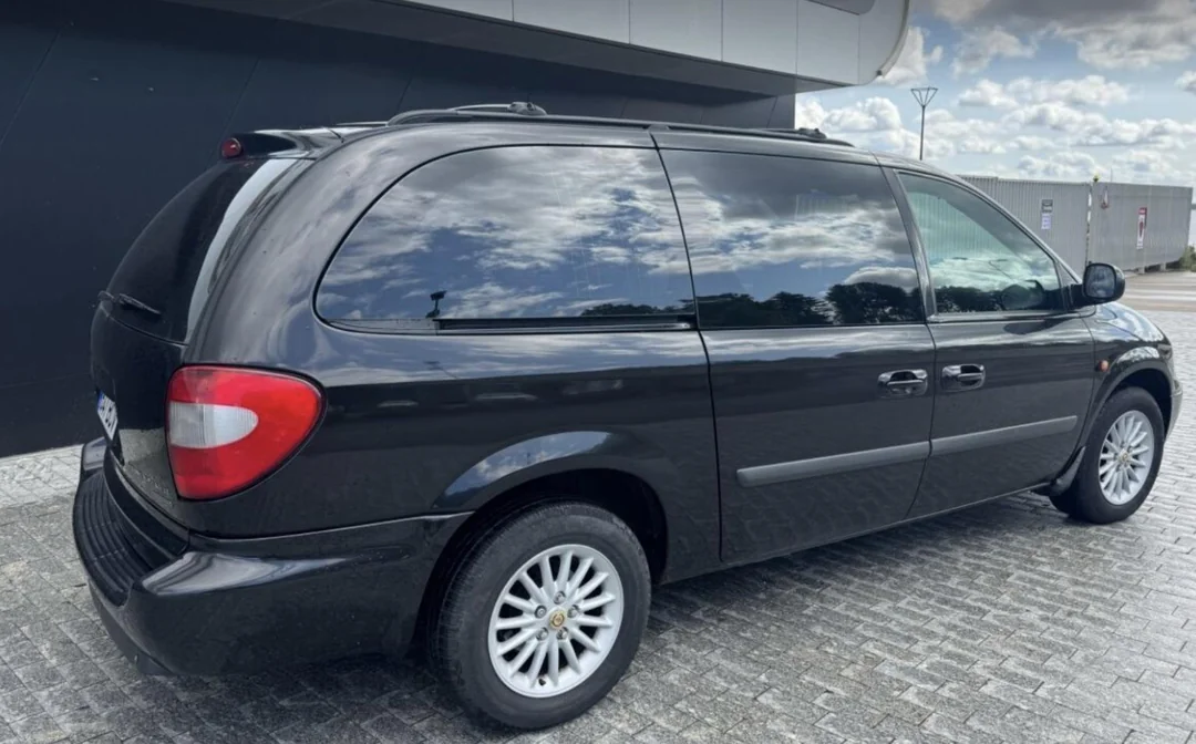 A black minivan is parked on a paved surface near a modern building, with reflections of clouds and sky visible on its windows and body.