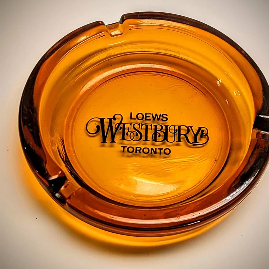 Amber-colored glass ashtray with the text “Loews Westbury Toronto” printed in black on the inside, shown against a white background.