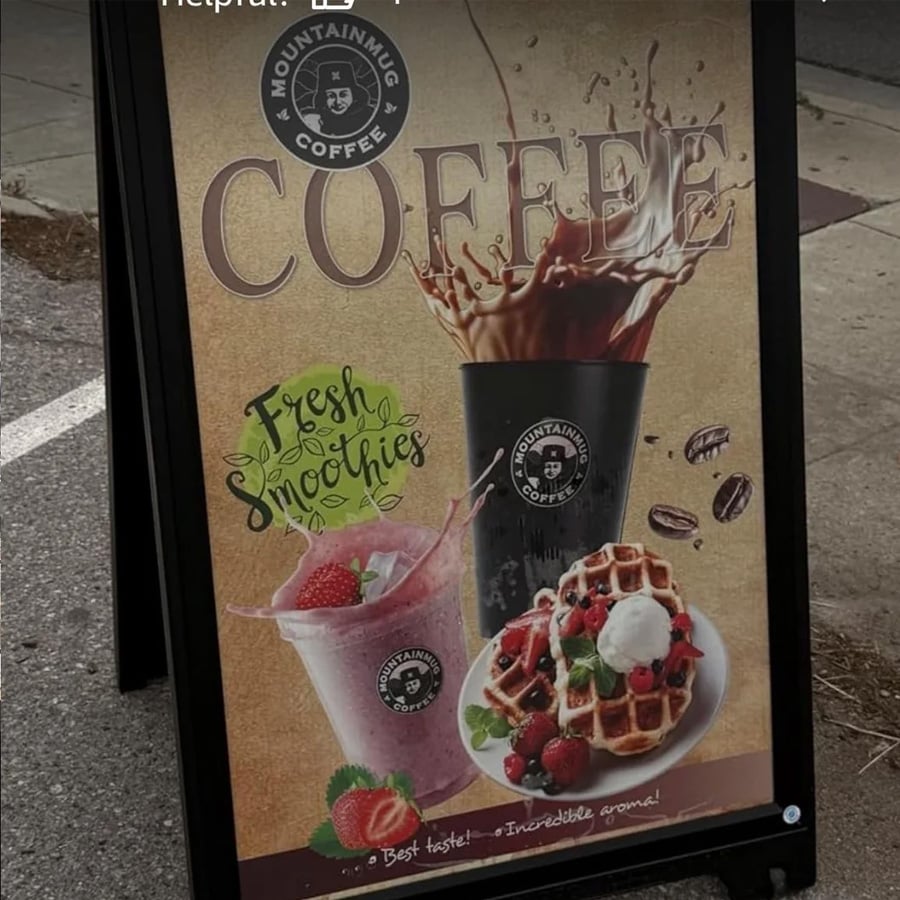 A sidewalk sign advertises Mountainhug Coffee, featuring splashing iced coffee, a pink smoothie with berries, and a plate of waffles with fruit. Text reads “Fresh Smoothies” and “Best taste! Incredible aroma!”