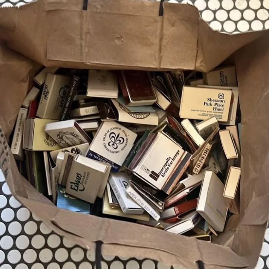 A paper bag filled with many matchbooks from various hotels and locations, sitting on a white tile floor with a black circle pattern.