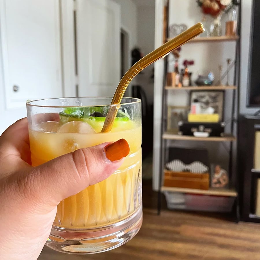A hand with orange nail polish holds a glass of orange drink with ice, lime, and a gold straw. A bookshelf and home decor are visible in the background.
