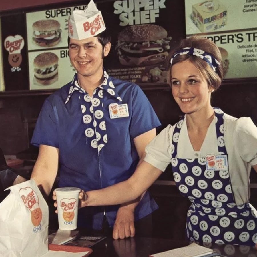 Two smiling fast food employees in vintage uniforms with smiley faces serve a drink and a bag to a customer at a Burger Chef counter, with menu boards visible in the background.