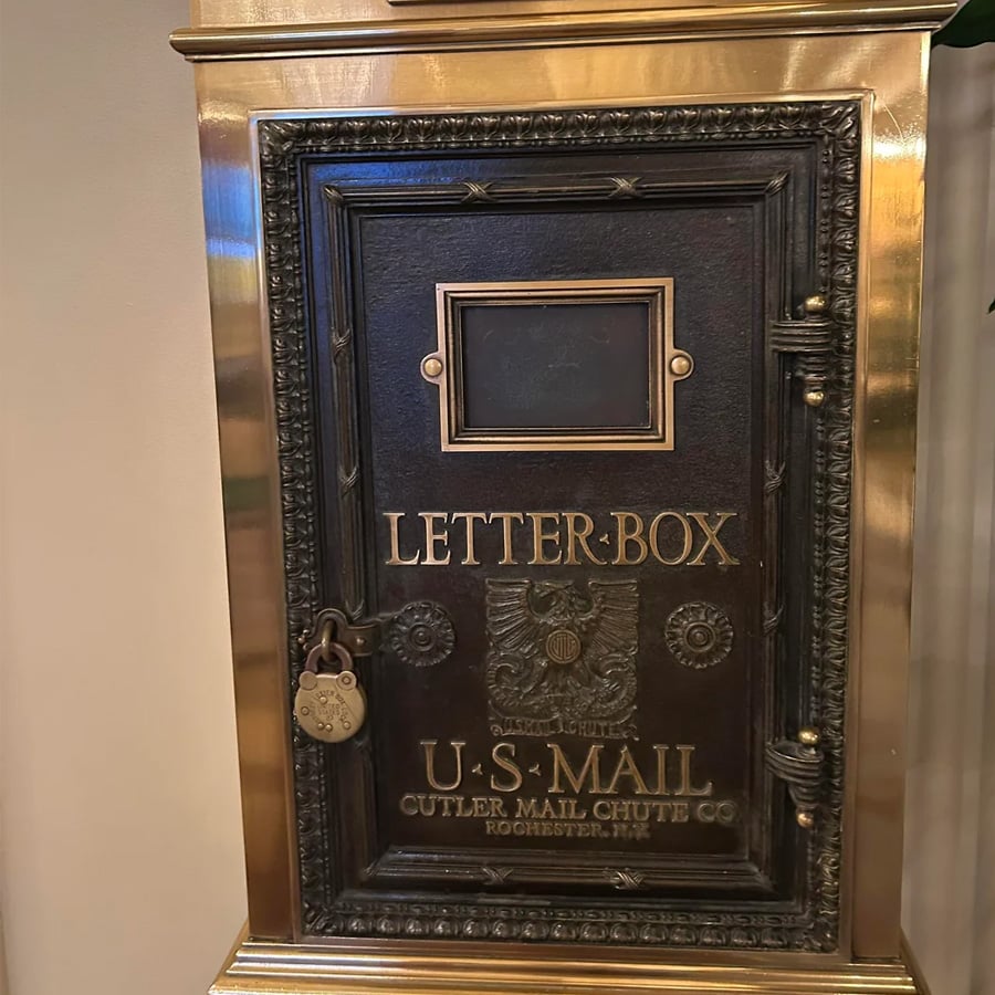 A vintage brass and black letterbox with ornate detailing, labeled “LETTER BOX U.S. MAIL” and secured with a padlock. Manufacturer inscription: “Cutler Mail Chute Co. Rochester, NY.”