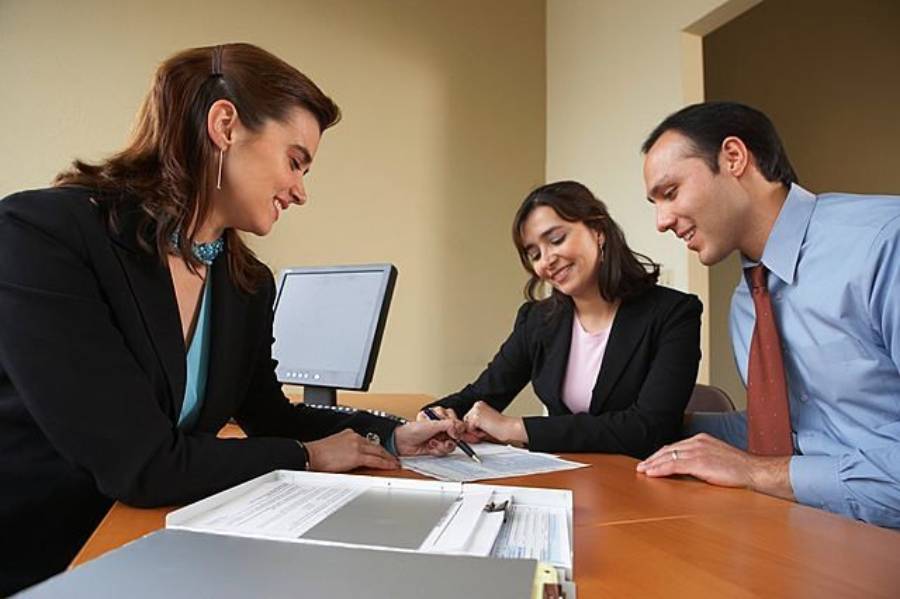 Three people in business attire sit at a desk, reviewing and signing documents together. A computer monitor and paperwork are visible on the desk. All appear focused and engaged in the discussion.