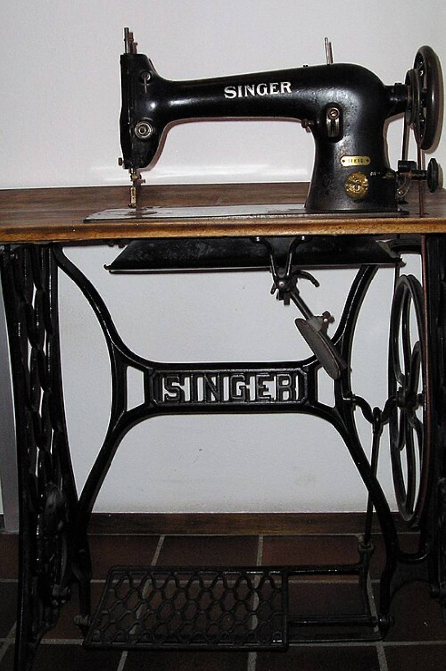 A vintage black Singer sewing machine with a metal treadle base, mounted on a wooden table. The word “SINGER” appears on both the machine and the base frame.