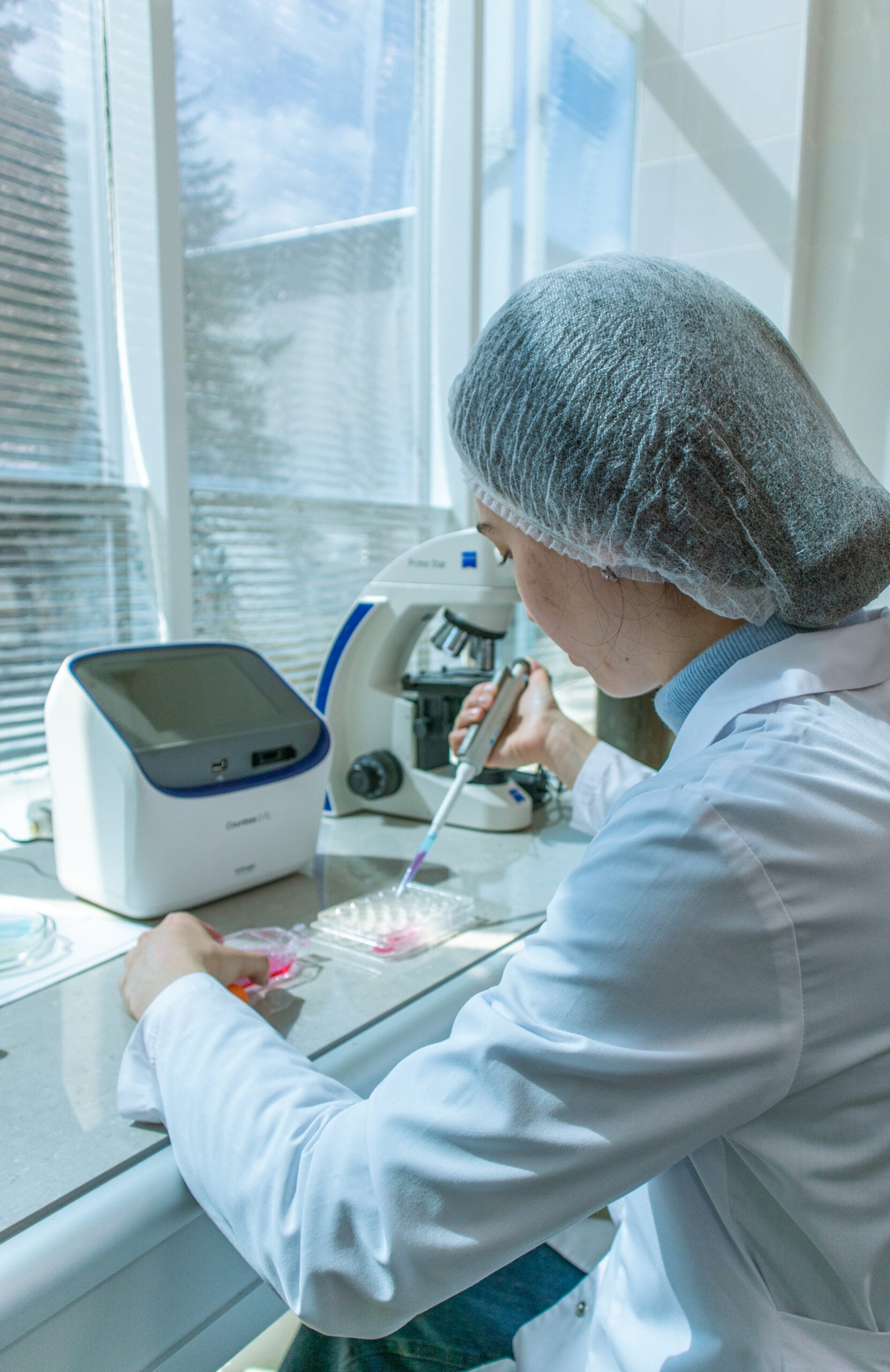 A scientist in a lab coat and hair net uses a pipette at a laboratory workstation near a window, with lab equipment including a microscope and analyzer nearby.