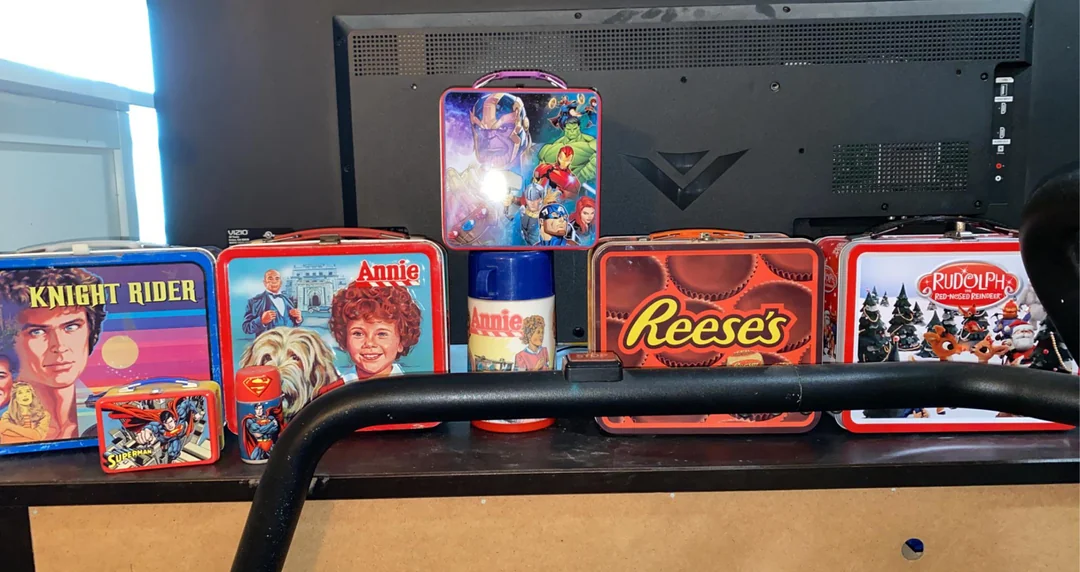 A collection of colorful vintage lunchboxes with themes like Knight Rider, Annie, superheroes, Reese’s, and Rudolph, displayed in front of a computer monitor.