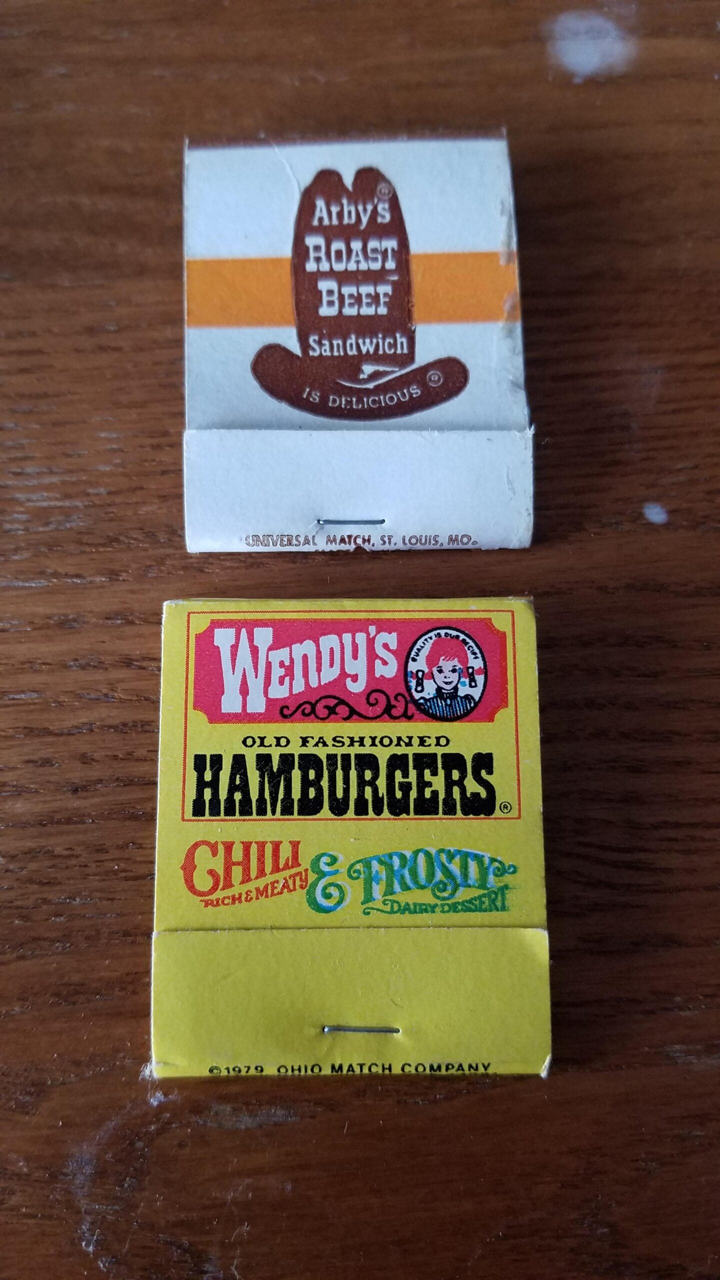Two vintage matchbooks on a wooden surface: one from Arby’s (white with brown logo) and one from Wendy’s (yellow, advertising hamburgers, chili, and Frosty dessert).