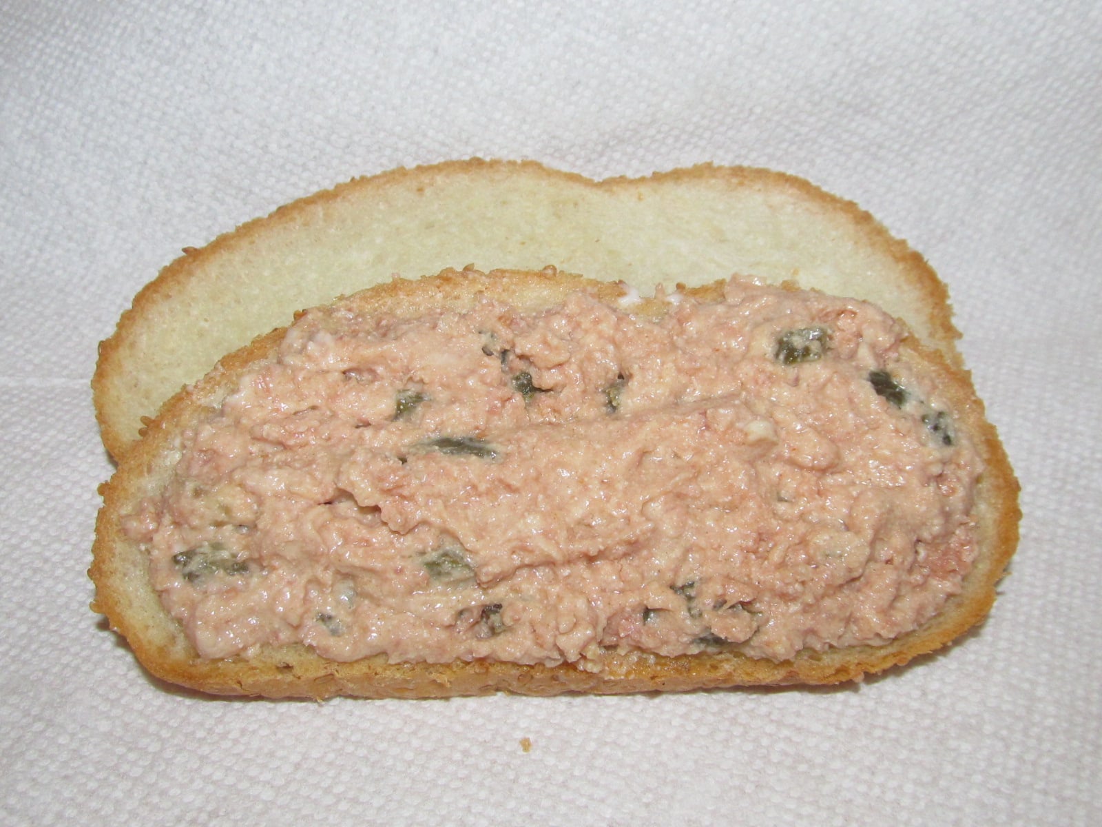 Two slices of white bread, one topped with a pinkish tuna or salmon salad mixture that contains small green bits, likely pickles or relish, placed on a white textured napkin.