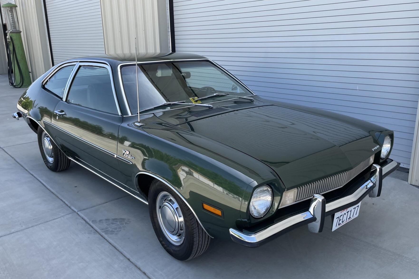 A dark green vintage Ford Pinto car is parked on a concrete driveway next to a corrugated metal building. The car has chrome trim, round headlights, and a classic 1970s design.