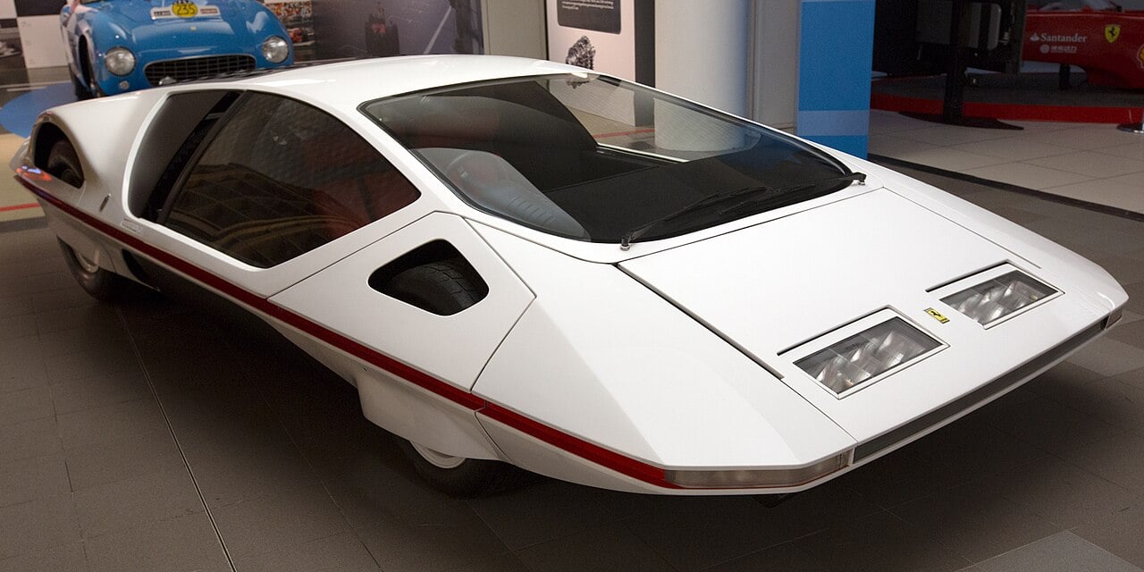 A futuristic, low-profile white concept car with sharp angles and large glass windows is displayed indoors. The car has rectangular headlights and minimal detailing, evoking a sleek, sci-fi aesthetic.