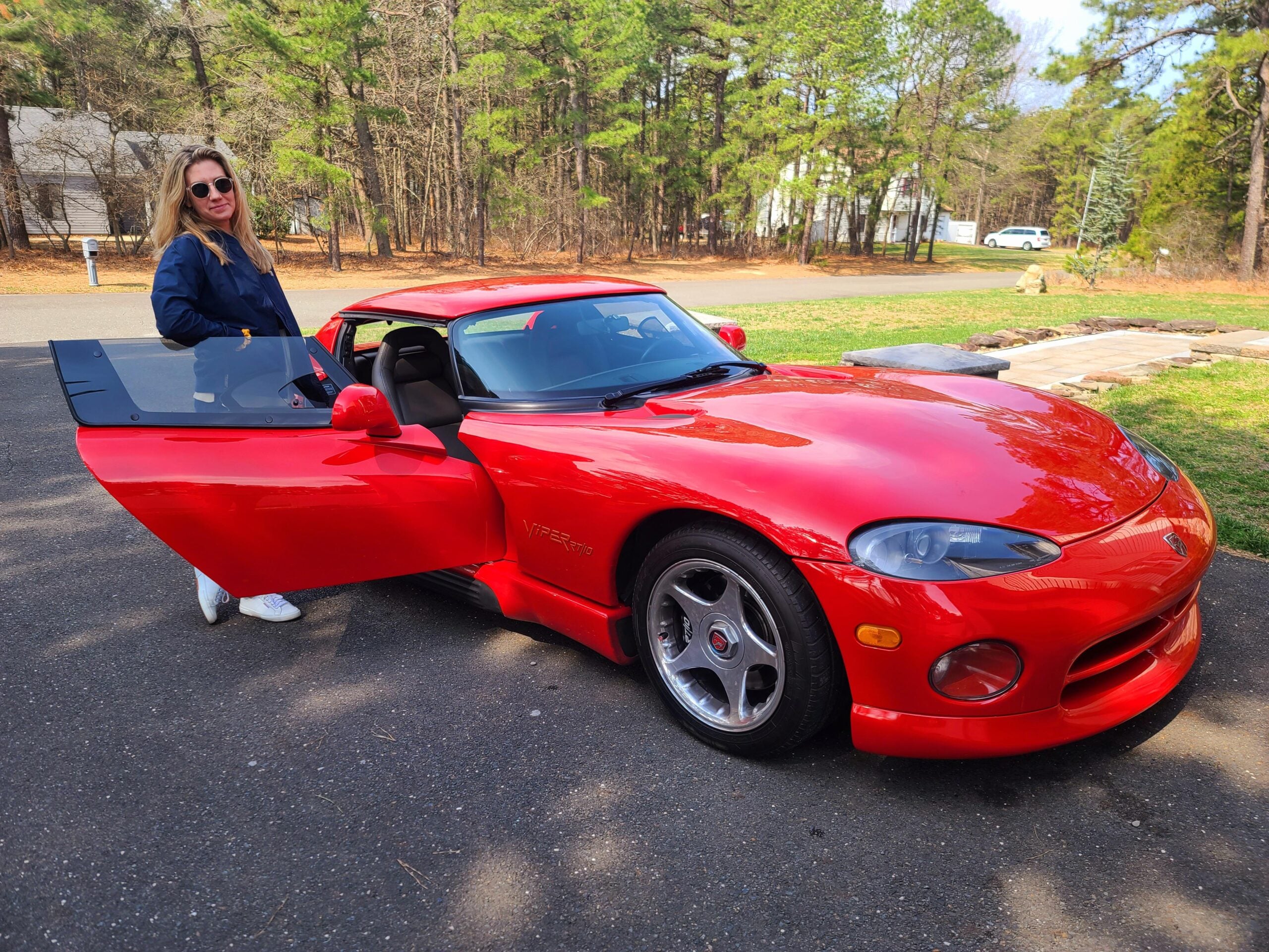 A woman wearing sunglasses stands next to a bright red sports car with the driver’s door open, parked on a paved driveway surrounded by trees.
