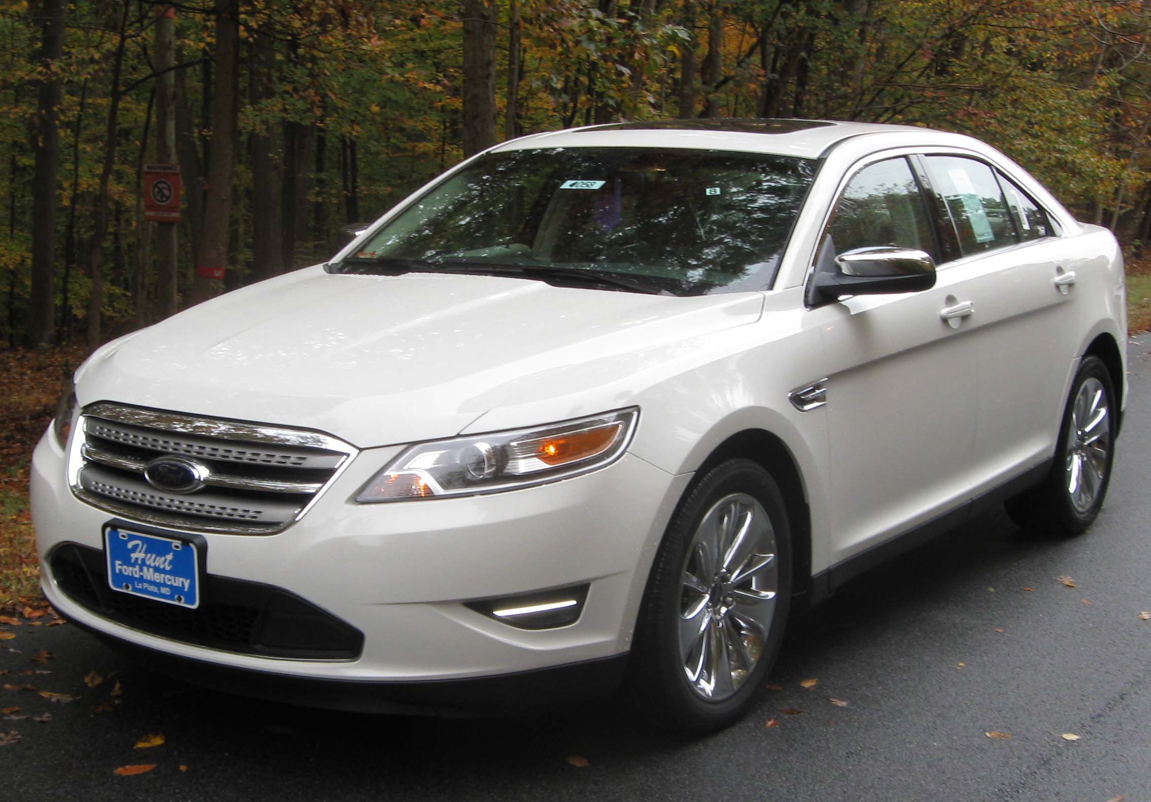 A white sedan parked on a wet road in front of autumn trees, with a blue dealership plate on the front bumper.