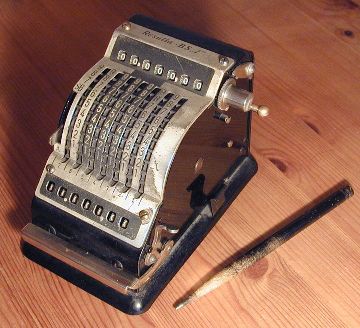 A vintage mechanical calculator with numbered dials and levers sits on a wooden table next to an old pen.