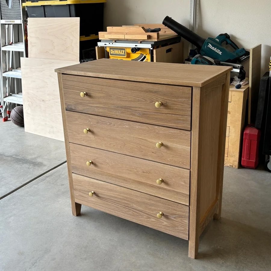 A wooden dresser with four drawers and gold knobs stands in a garage, surrounded by tools and workshop equipment.