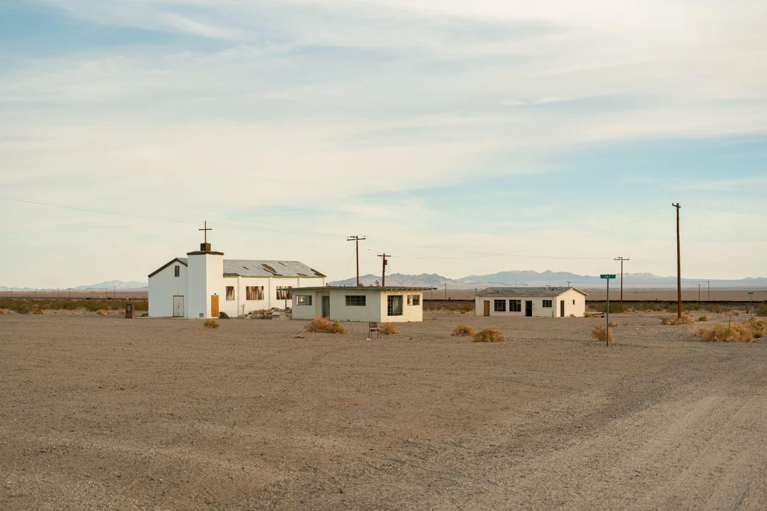 A small white church with a cross and two nearby buildings stand in a barren, gravel desert landscape under a partly cloudy sky, with distant mountains in the background.