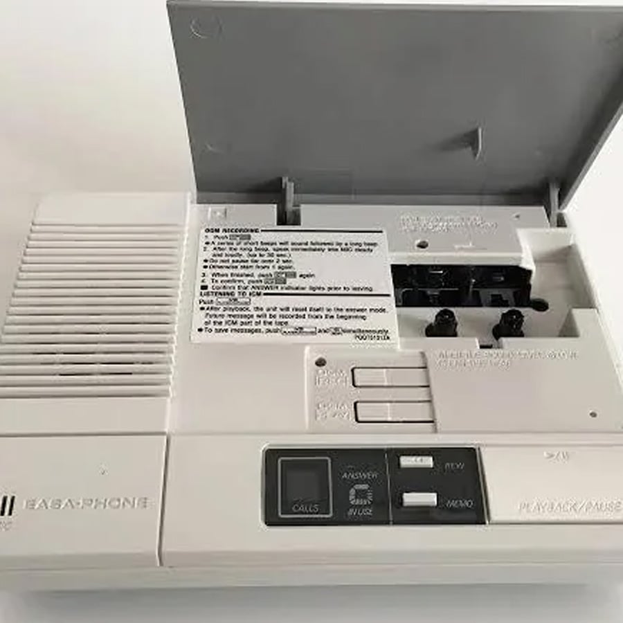 A white vintage answering machine with its cassette compartment open, buttons for play, rewind, and answer functions visible, and an instruction label inside the lid.