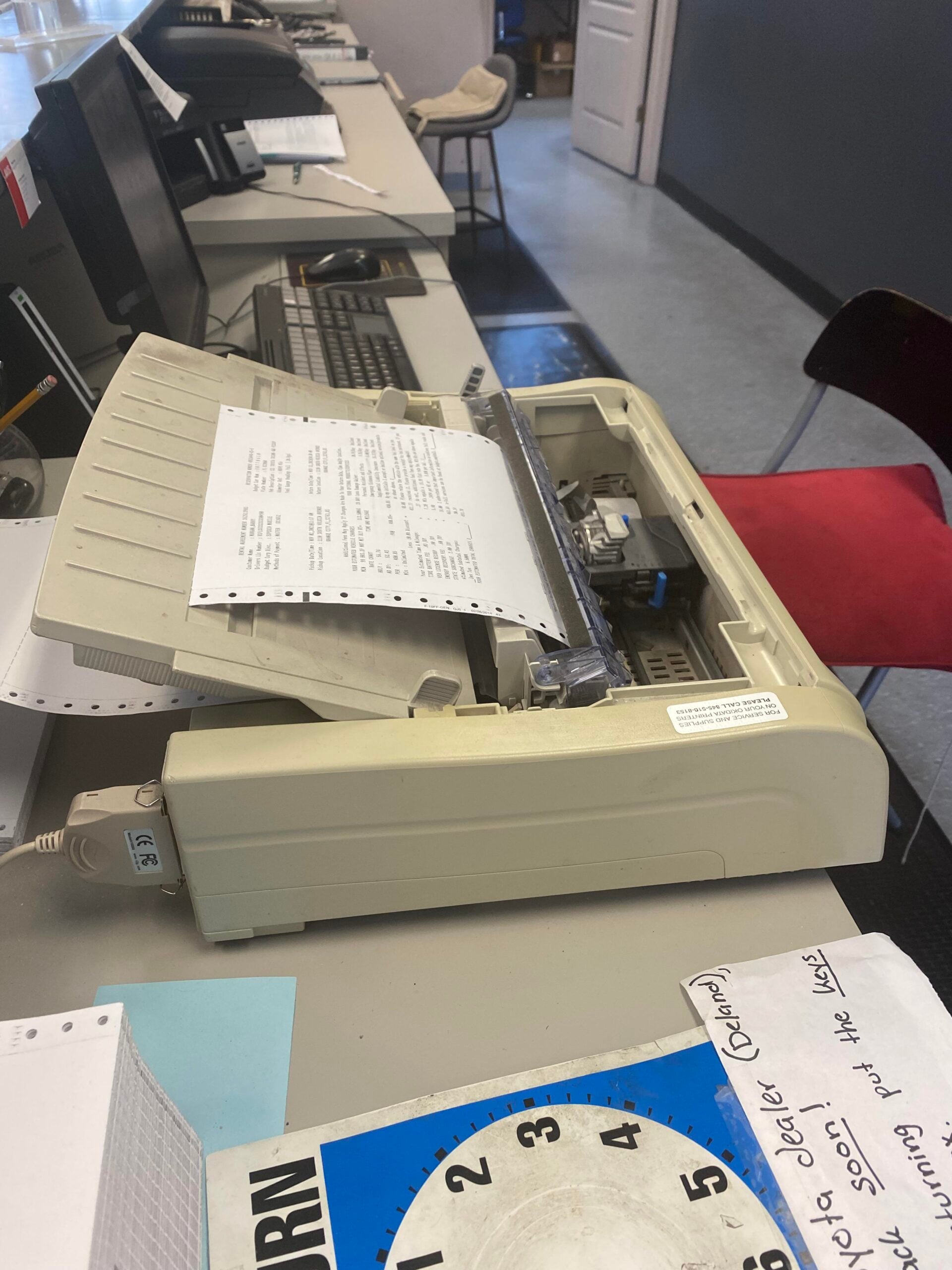 A dot matrix printer is printing a perforated paper form on a desk with computers. A red chair is nearby, and a blue and white circular form is partially visible in the foreground.