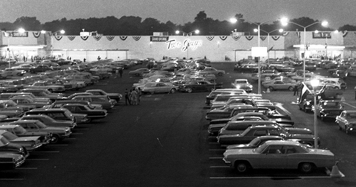 Black and white photo of a crowded parking lot at night, filled with vintage cars. In the background, a large building displays the sign “Two Guys” and patriotic banners. Tall lights illuminate the scene.