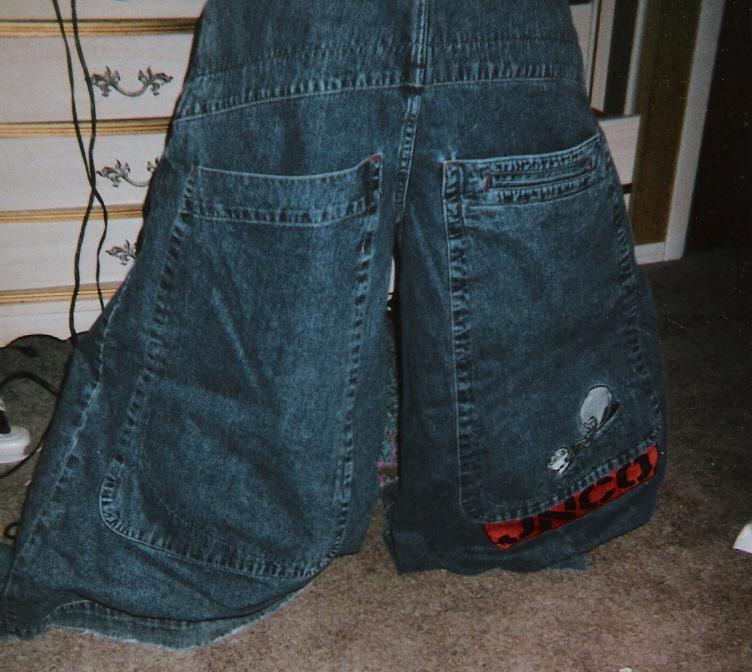 A person wearing oversized blue jeans with very large back pockets, one pocket featuring a skull design and red accent, standing indoors near a dresser.