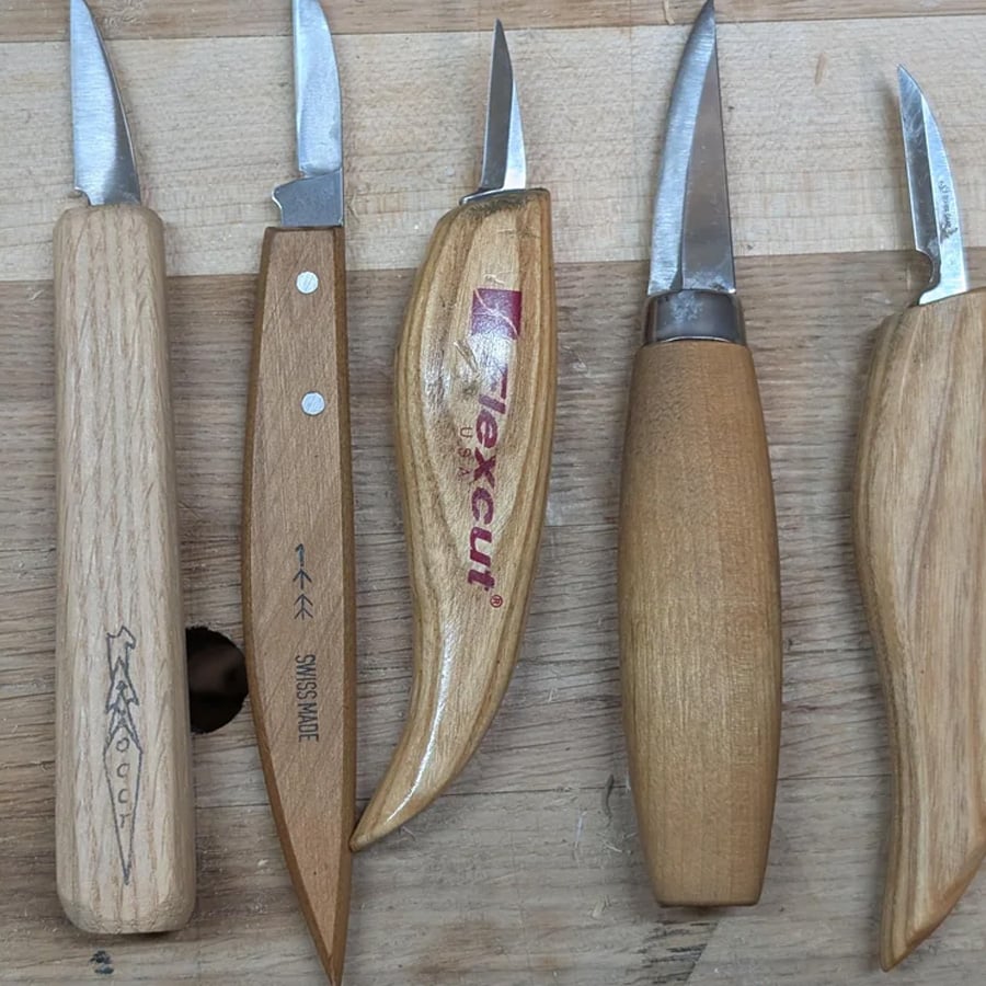 Five wood carving knives with wooden handles are arranged side by side on a wooden surface. Each knife has a different blade shape and handle design, some with brand markings visible.