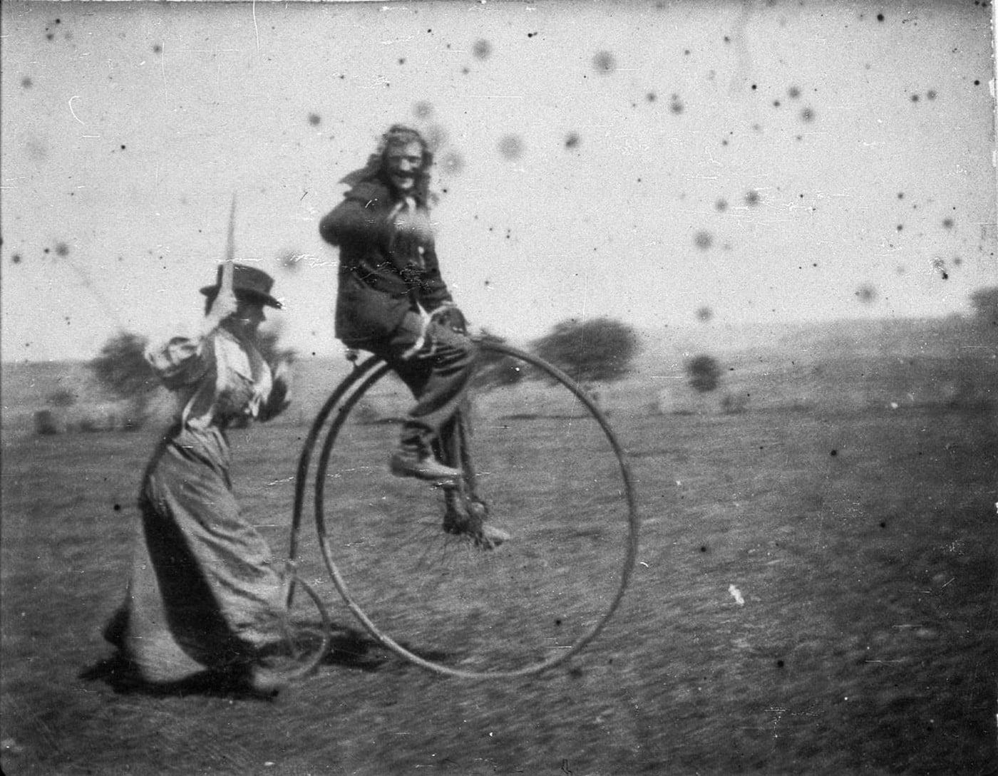 A person in old-fashioned clothing rides a vintage penny-farthing bicycle while another person, also in period dress, runs behind them outdoors on a grassy field. The photo is black and white and shows some age spots.