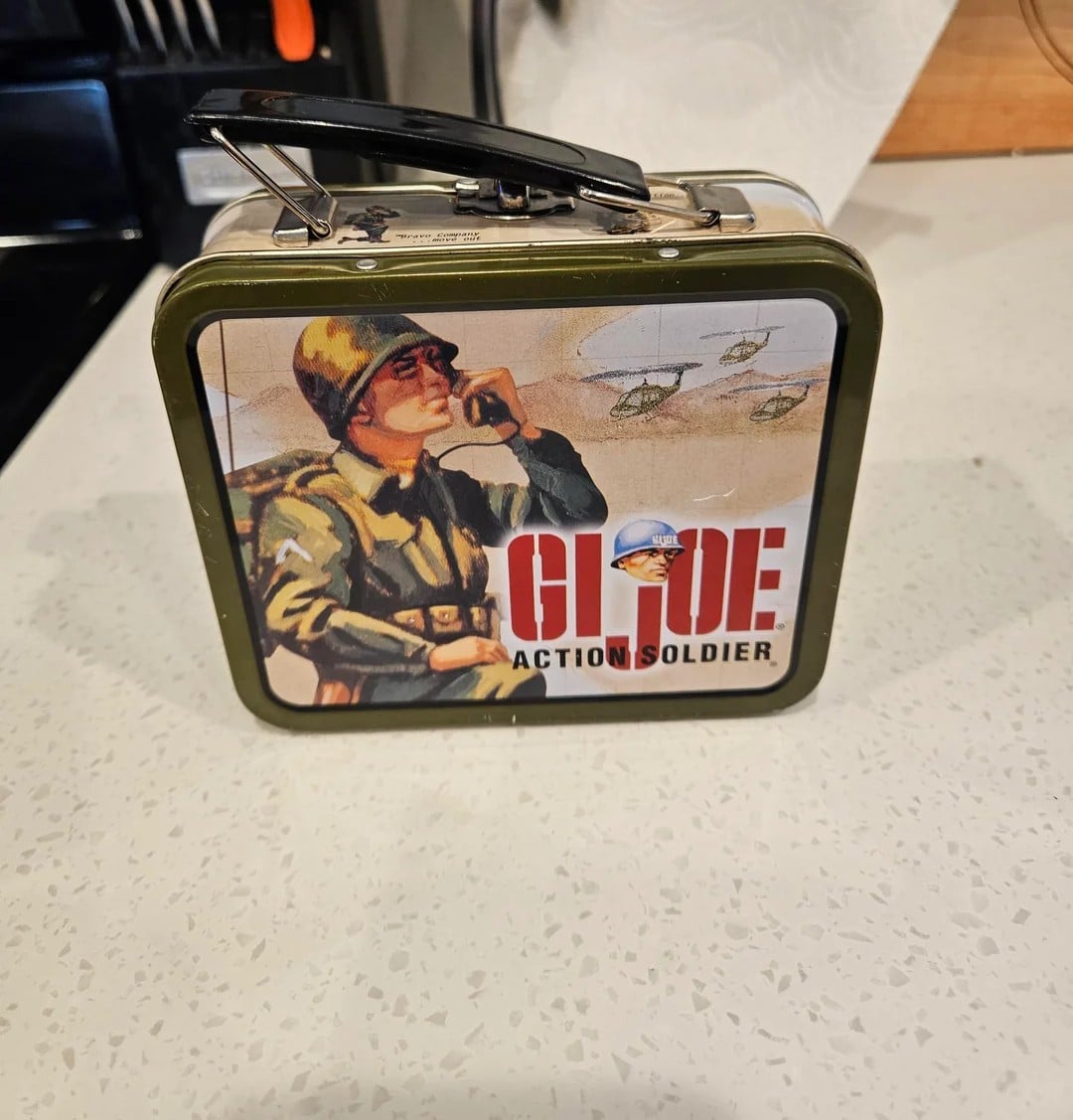 A metal lunchbox featuring vintage-style G.I. Joe artwork of a soldier with a walkie-talkie, helicopters in the background, and the text “G.I. Joe Action Soldier” on a kitchen countertop.