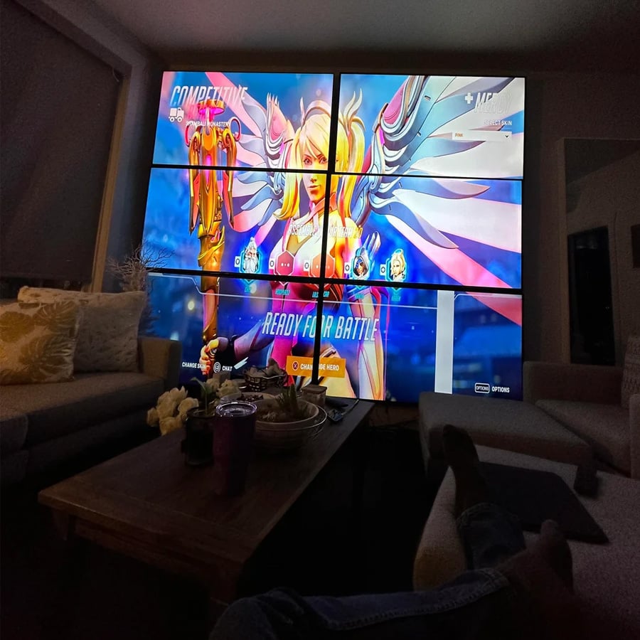 A large TV wall displays the character selection screen for Mercy in Overwatch in a dimly lit living room, with couches, a table, snacks, and someone’s feet visible in the foreground.