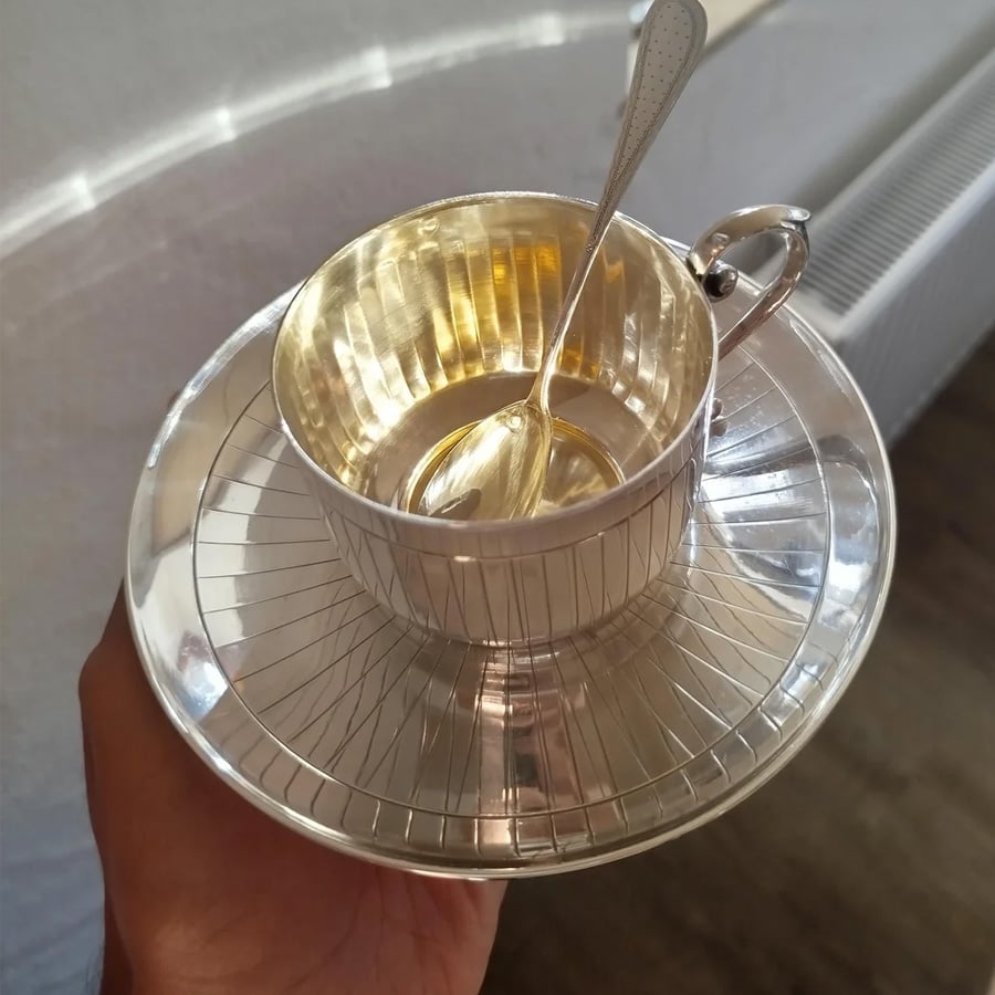 A hand holds a shiny silver teacup with a matching saucer and a spoon inside. The reflective set features fine line detailing and catches the light.