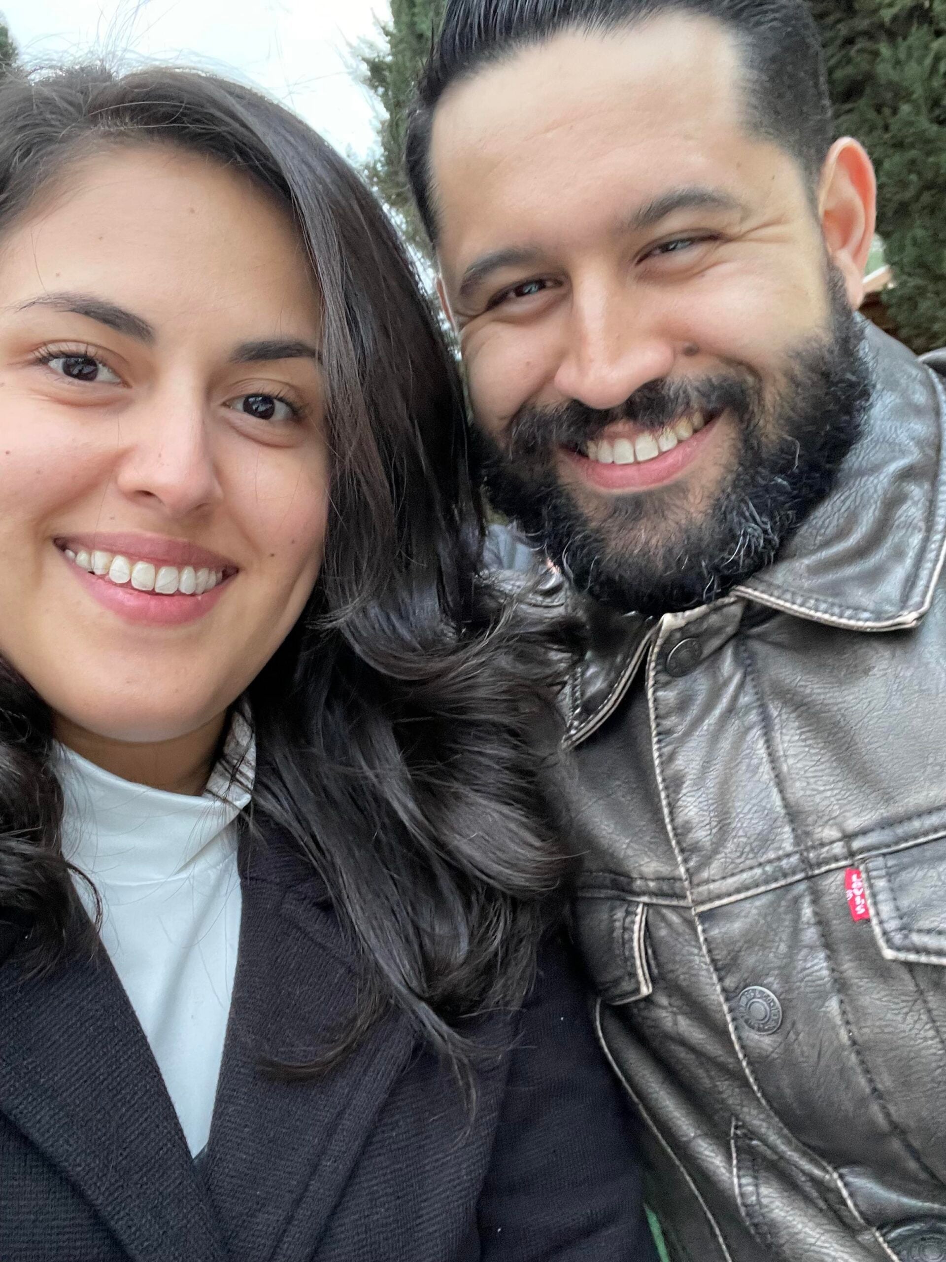 Two people smiling and posing closely together outdoors, both with dark hair. The person on the left wears a black coat, and the person on the right wears a shiny metallic jacket with a beard. Trees are blurred in the background.