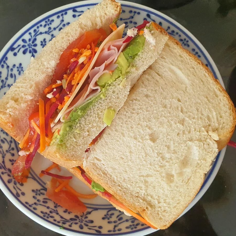 A sandwich on white bread filled with slices of turkey, avocado, shredded carrots, red cabbage, and tomato, served on a blue and white patterned plate.