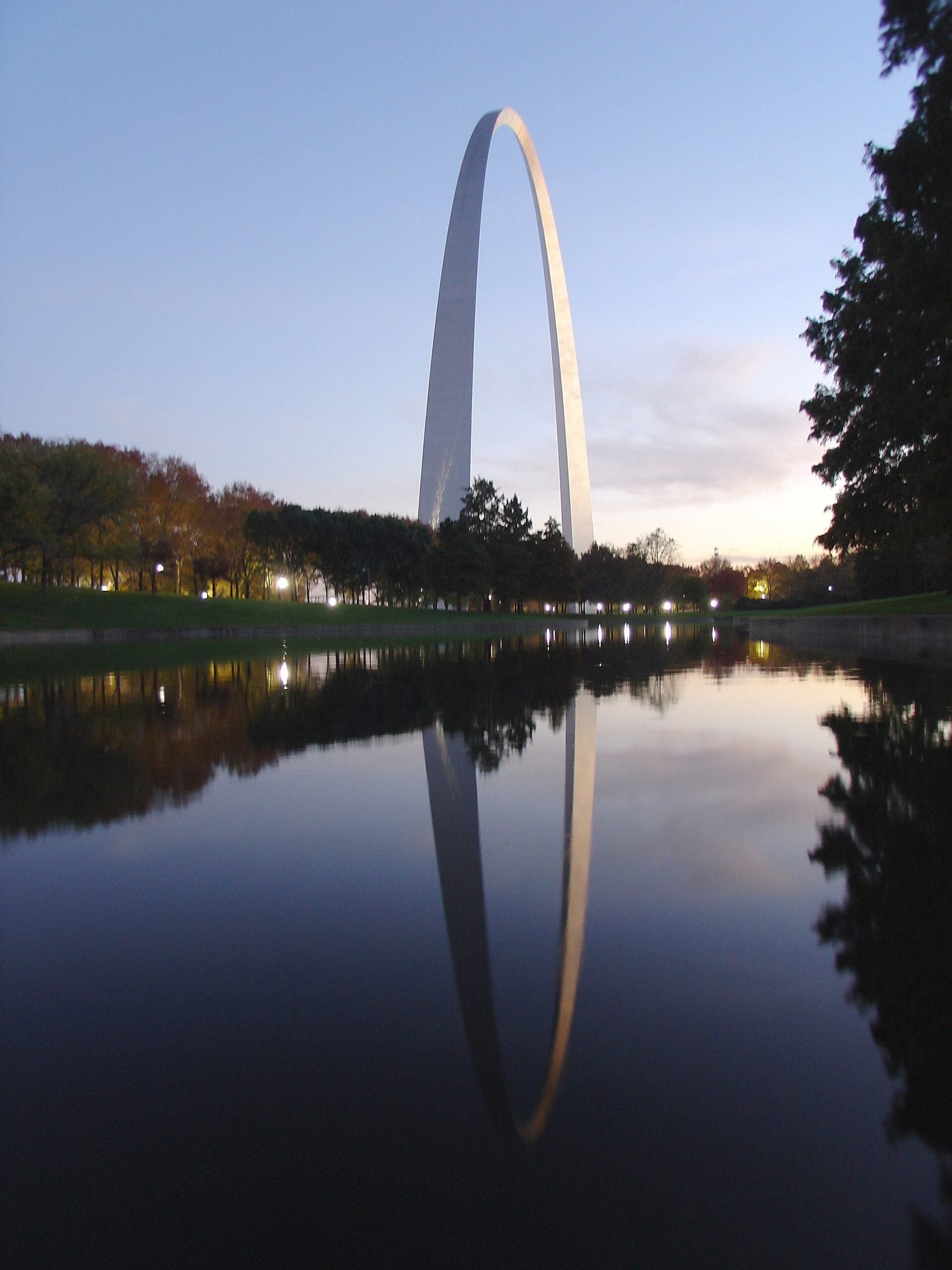 The Gateway Arch in St. Louis is reflected in a calm body of water at dusk, with trees lining both sides and the sky fading from blue to light near the horizon.