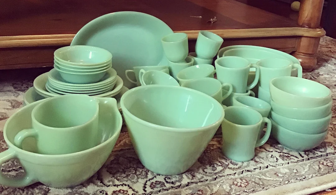 A collection of vintage green glass dishes, including bowls, plates, cups, and pitchers, arranged on a patterned rug in front of a wooden table.