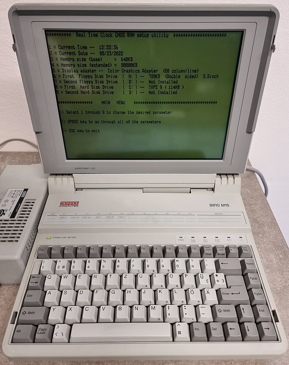 A vintage Siemens Nixdorf laptop, the BSO M10, displays a green-on-black BIOS setup screen. The laptop sits on a table with its keyboard visible and a connected power adapter on the left side.