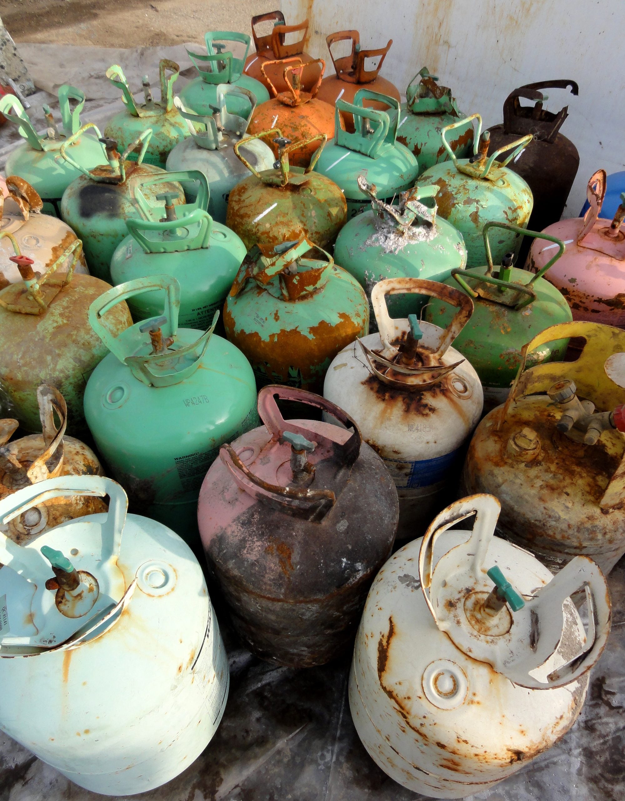 A group of old, rusted gas cylinders in various colors, including green, white, and brown, are clustered together outdoors on a concrete surface.